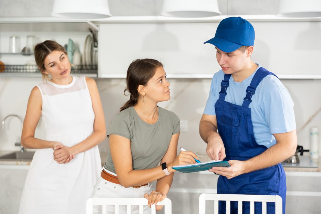 A workman in overalls and cap showing a clipboard to two women in a kitchen