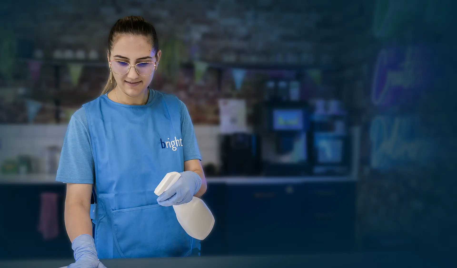 A cleaner in branded apron holding a sponge and spray bottle in a cafe-style setting with hanging bunting