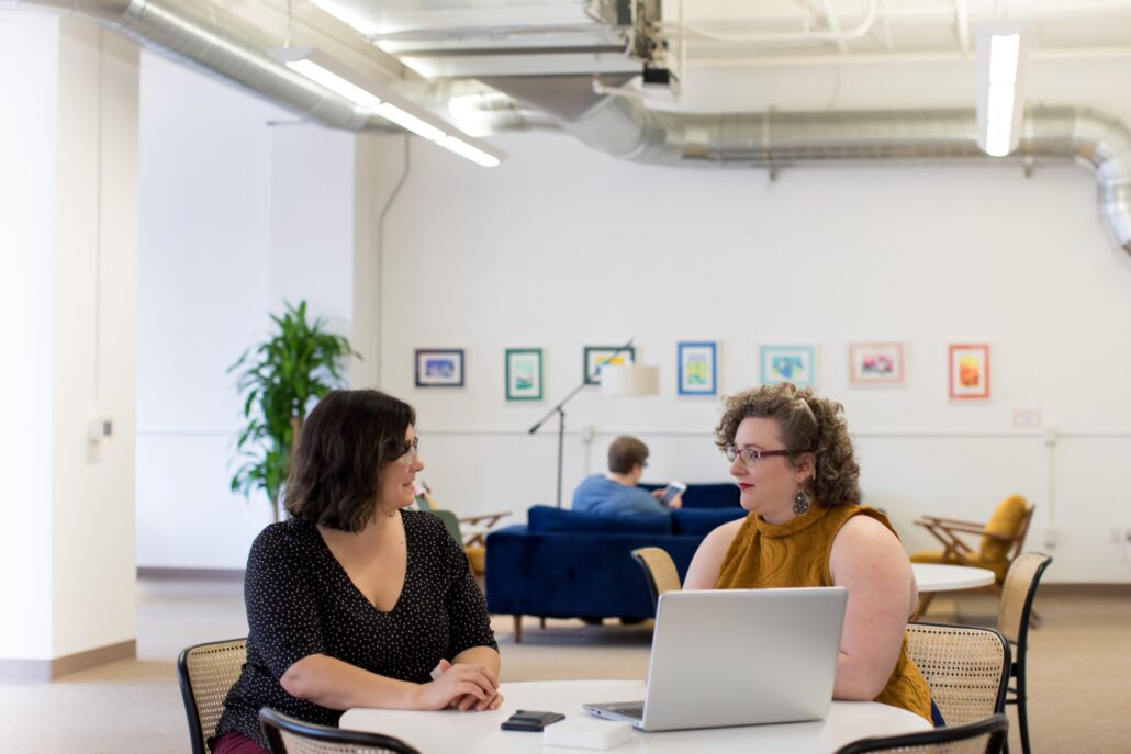 Two women chatting at a table with a laptop in an open-plan office with exposed ceiling ducts