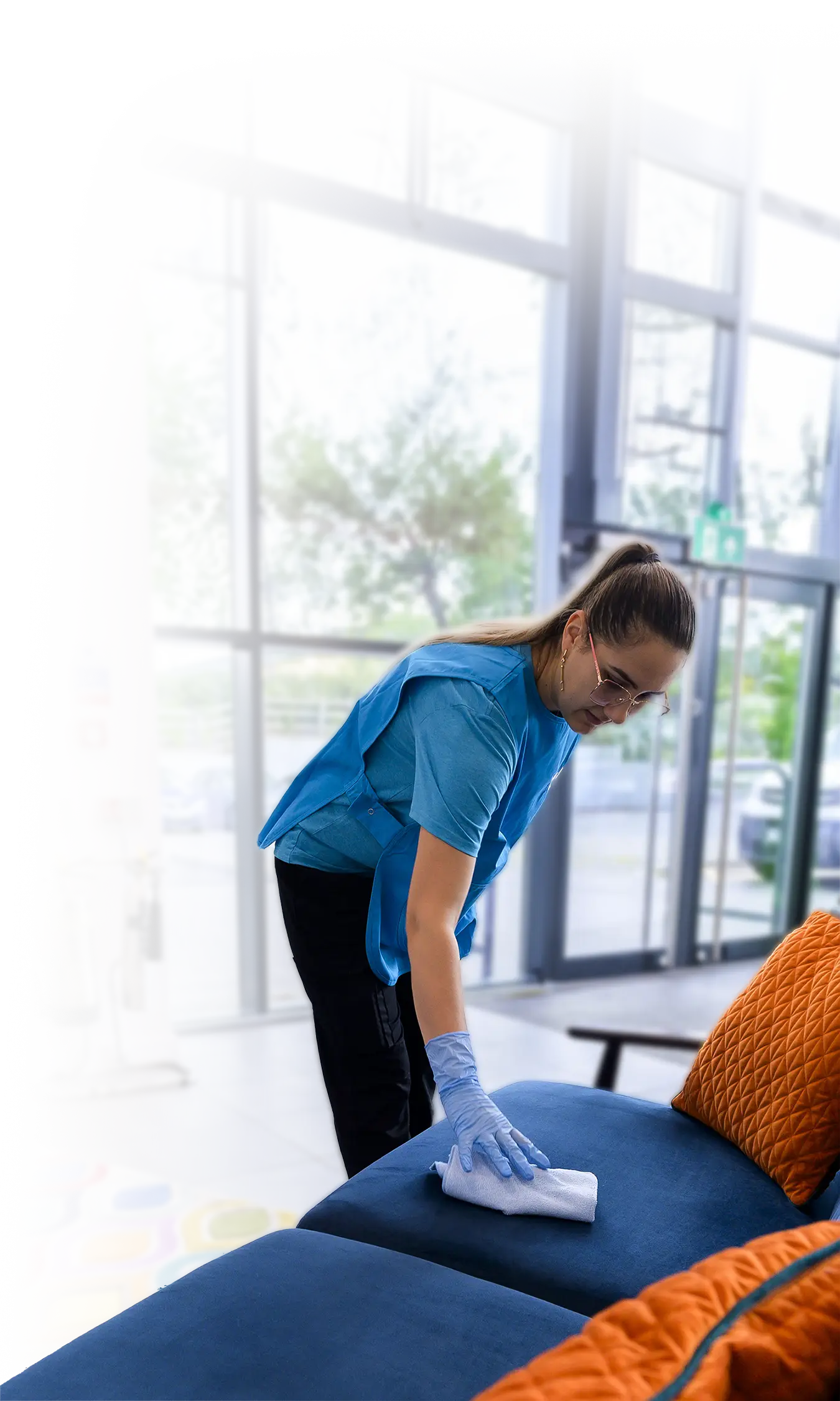 A cleaner in branded polo shirt wiping a blue sofa with a cloth in a glass-fronted lobby