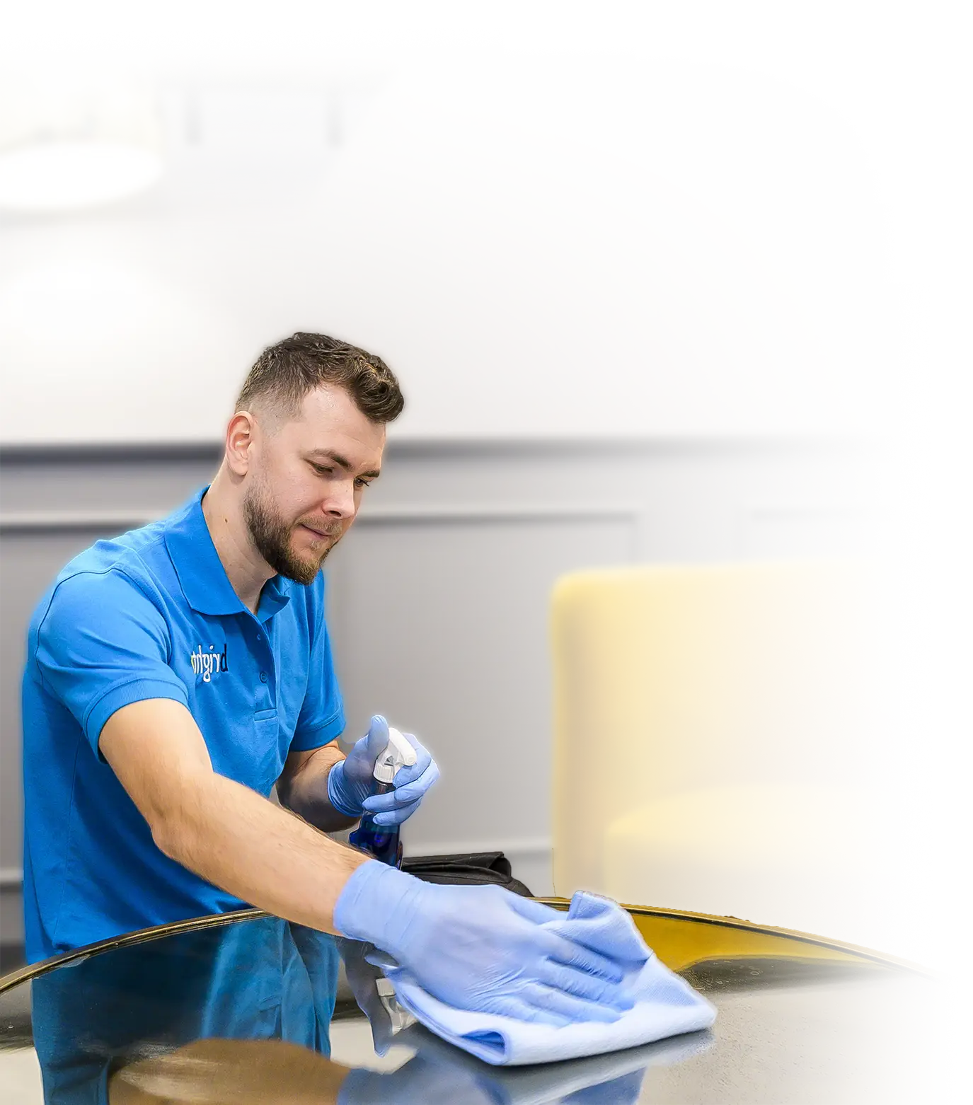 A cleaner in a branded polo spraying and wiping a glossy round table with a blue cloth