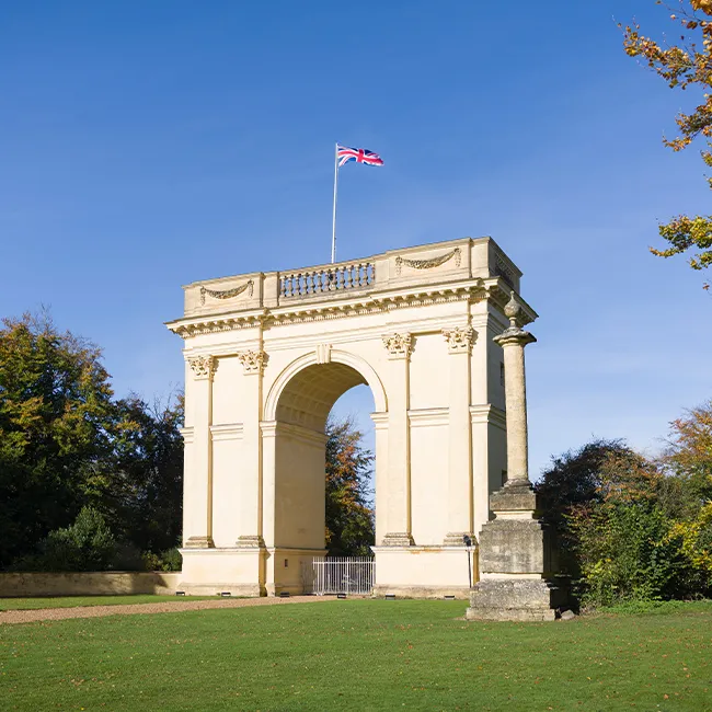 A stone triumphal arch with a union jack flag flying, set in parkland with trees and lawn