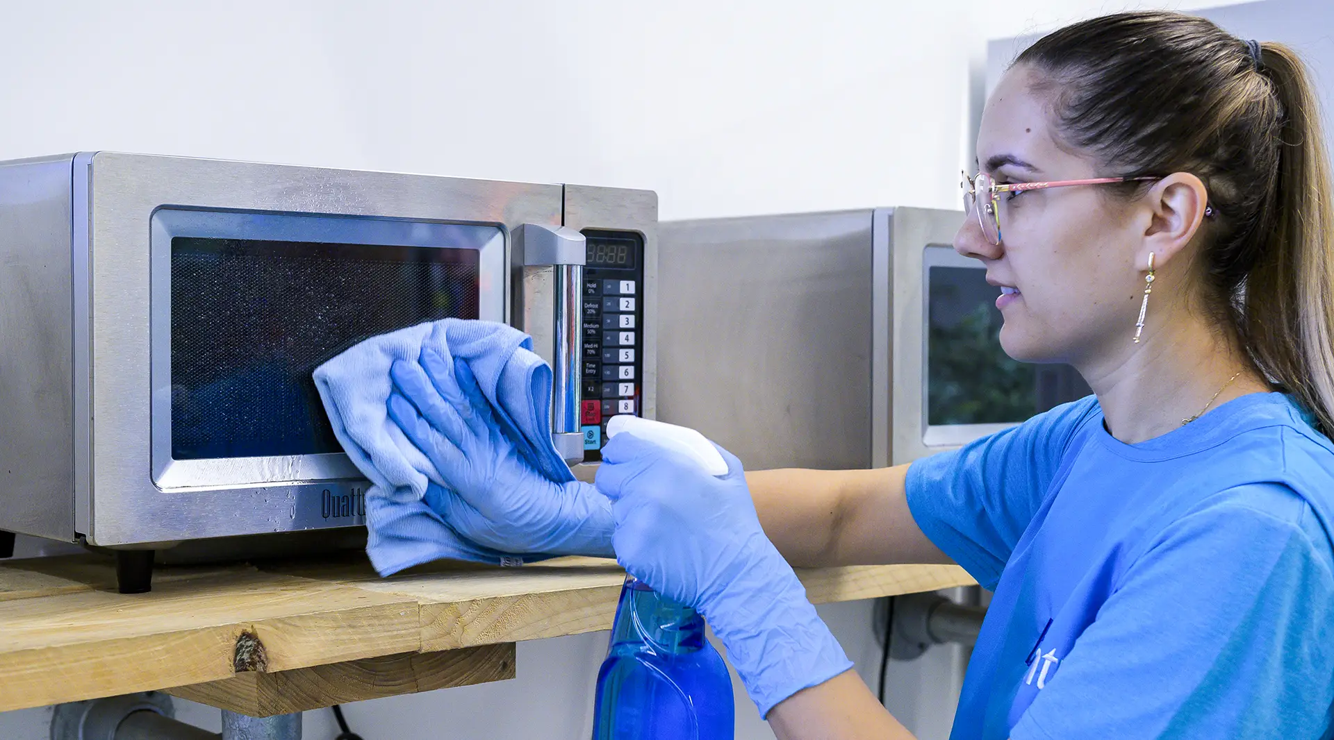 A cleaner in branded blue polo wiping a stainless steel commercial microwave with a cloth and spray