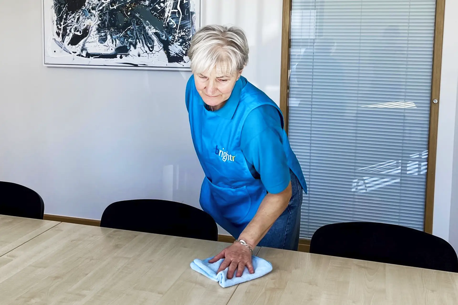 An older woman in a branded tunic wiping a long table in an office with abstract artwork on the wall