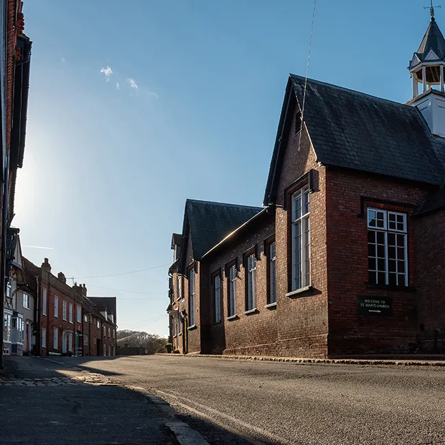 A brick building with a sloped roof and small tower on a quiet street at low sun, sign mounted on the wall