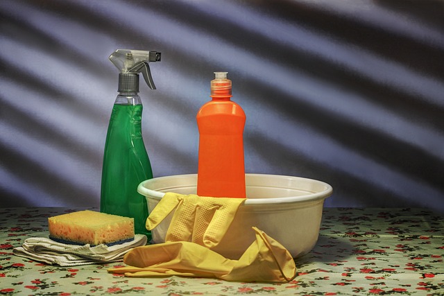 A green spray bottle, orange bottle, sponge, cloth and yellow rubber gloves beside a white bowl