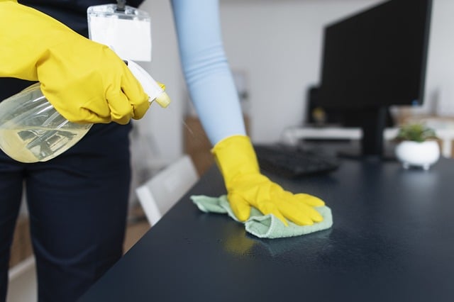 A gloved hand wiping a dark desk with a cloth and spray bottle, monitor in background
