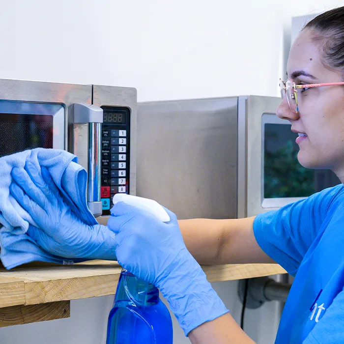 A cleaner in branded polo and blue gloves wiping the handle of a stainless steel microwave with a cloth