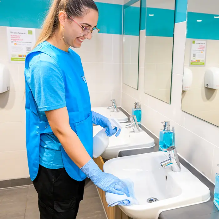 A cleaner in a branded tunic wiping washroom sinks with a spray bottle and blue gloves