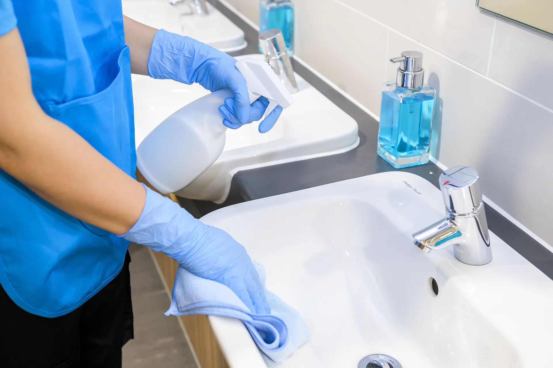 Gloved hands wiping a bathroom sink with a blue cloth and spray bottle, soap dispensers on the counter