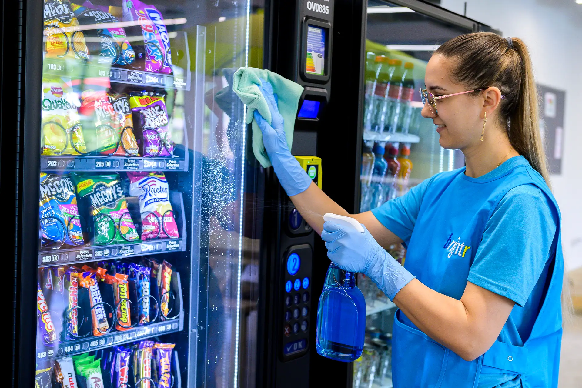 A cleaner in branded polo wiping the glass front of a vending machine stocked with snacks and drinks