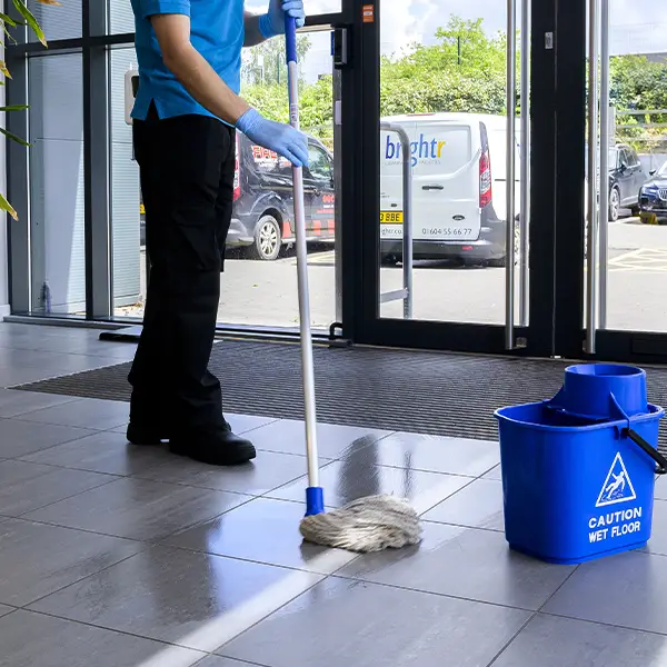 A cleaner mopping a tiled entrance floor beside a wet floor bucket, a branded van outside