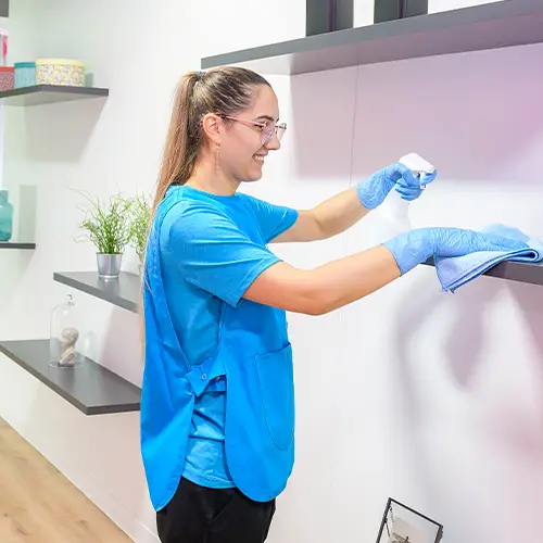 A cleaner in branded blue tunic and blue gloves spraying and wiping a dark wall-mounted shelf
