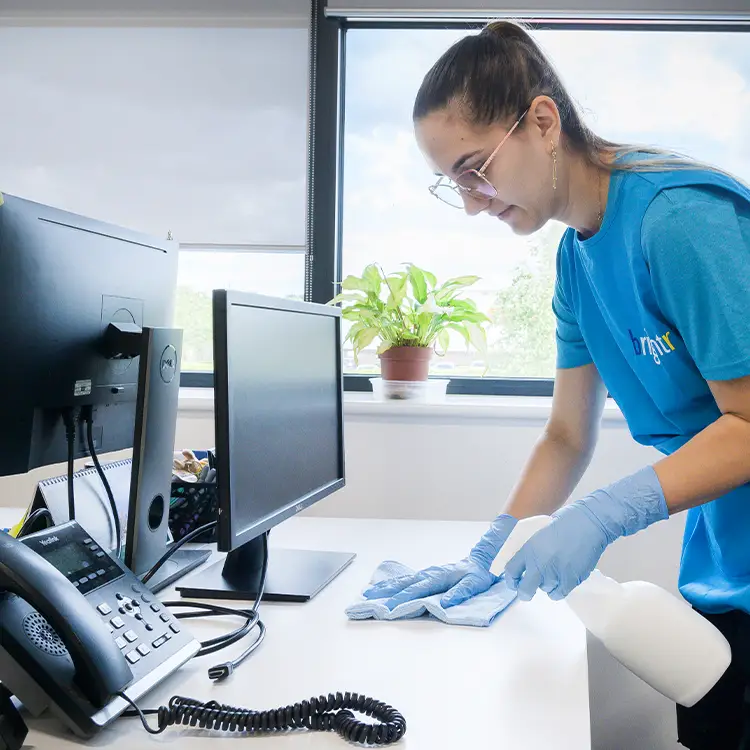 A cleaner in branded polo shirt wiping an office desk beside computer monitors, wearing blue gloves