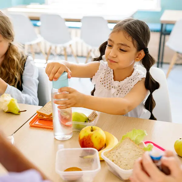 Children at a table with lunchboxes and fruit, a girl opening a bottle of water