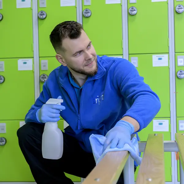 A cleaner in branded blue fleece wiping a wooden bench beside green lockers, holding a spray bottle
