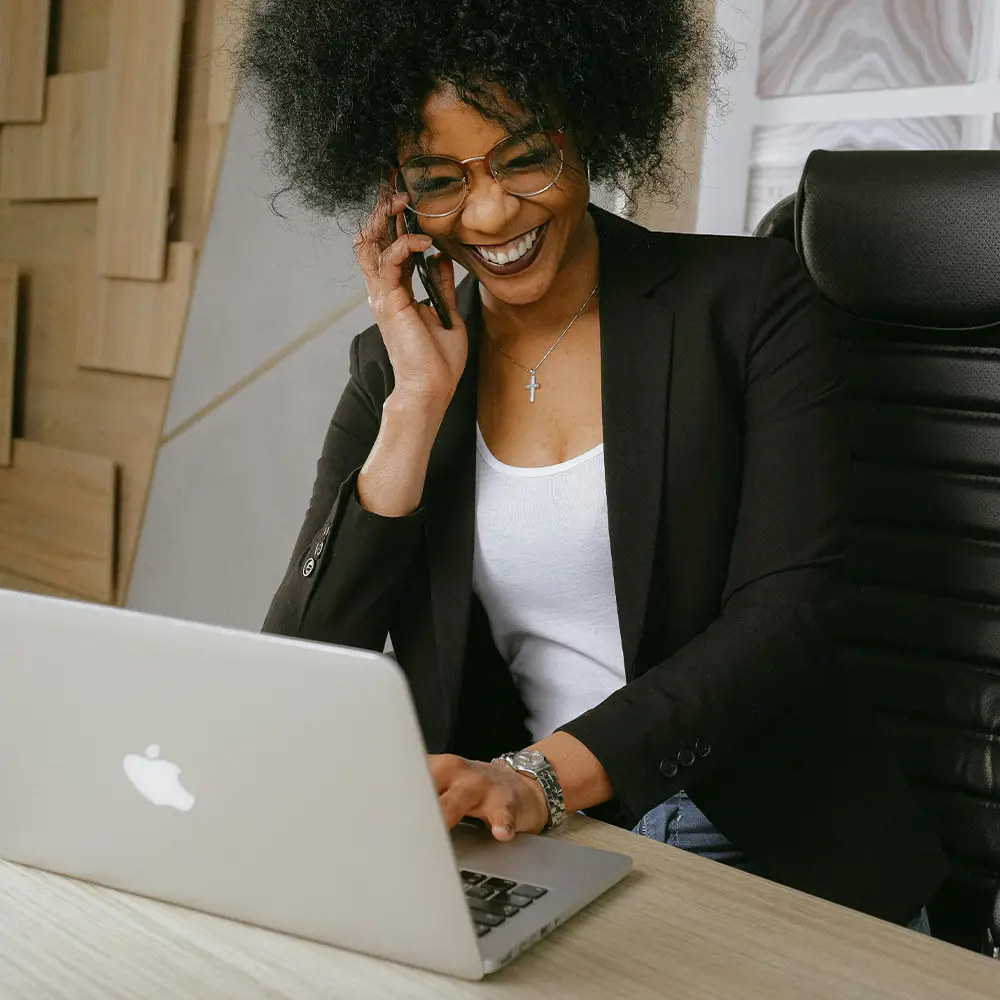 A woman in a blazer smiling on her phone while typing on a laptop at a desk