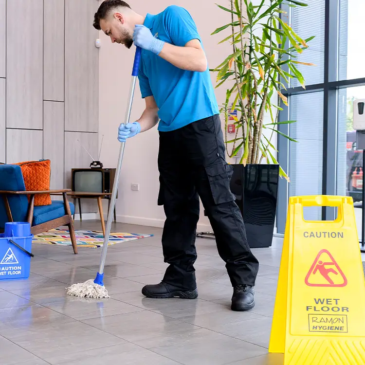 A cleaner in branded polo mopping a tiled floor beside a wet floor caution sign and mop bucket