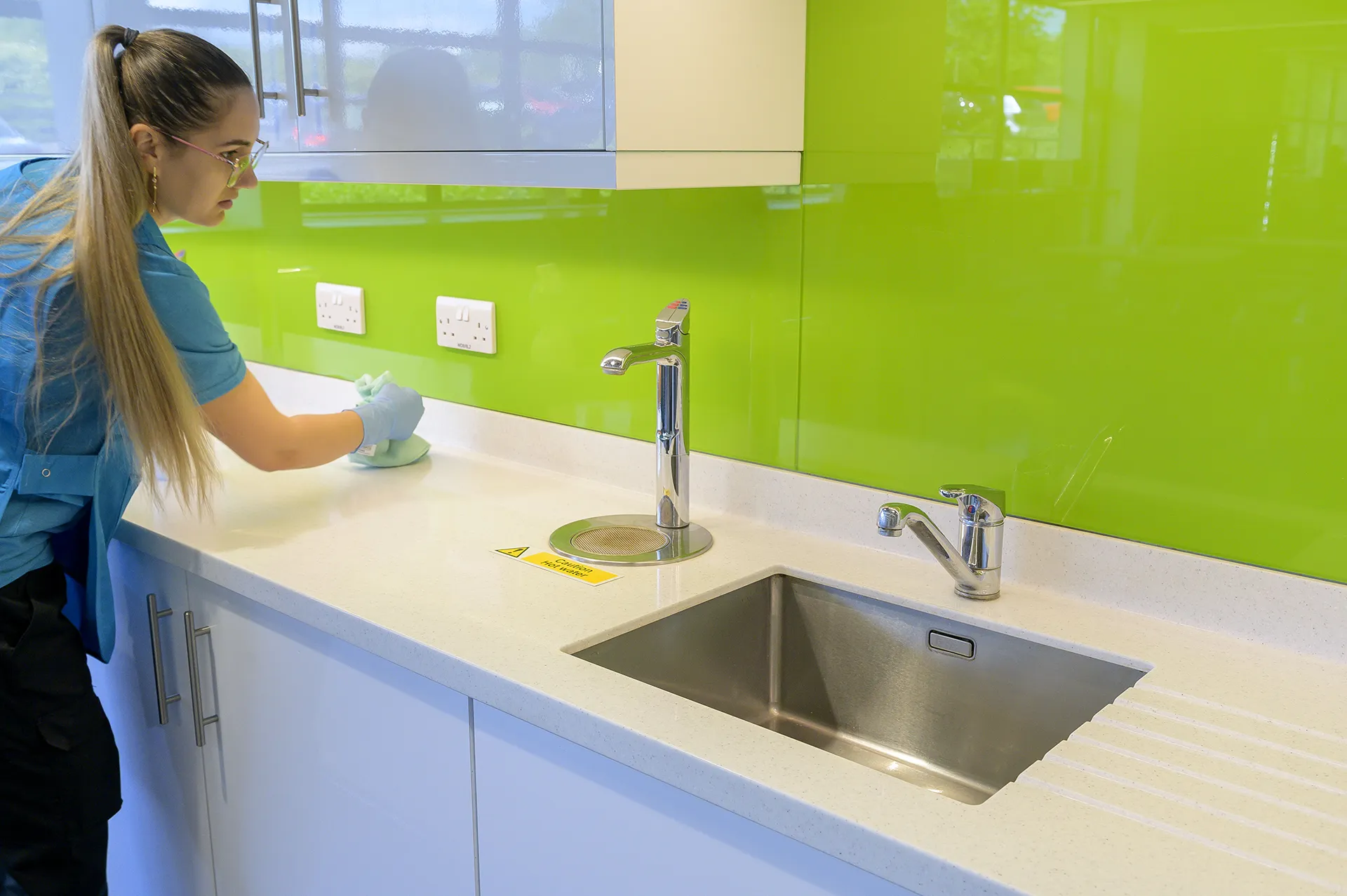 A cleaner in a branded blue polo wiping a counter beside a stainless sink, a 'Caution Hot Water' sign nearby