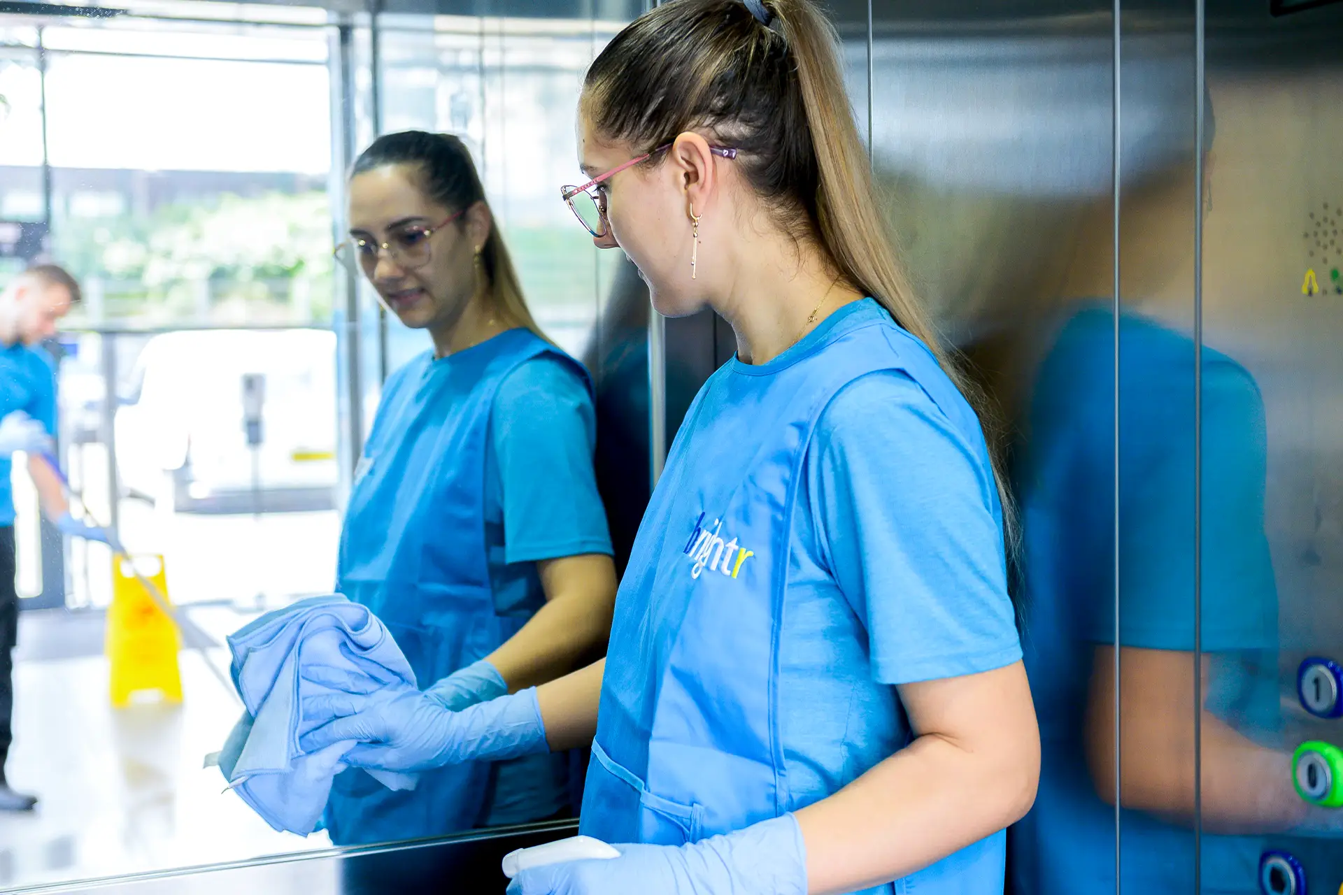 Two cleaners in branded uniforms cleaning a mirrored lift panel, a mop and wet floor sign reflected behind them