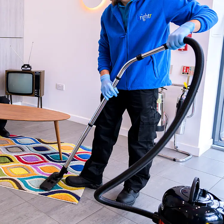 A cleaner in branded fleece and blue gloves vacuuming a patterned rug with a cylinder vacuum