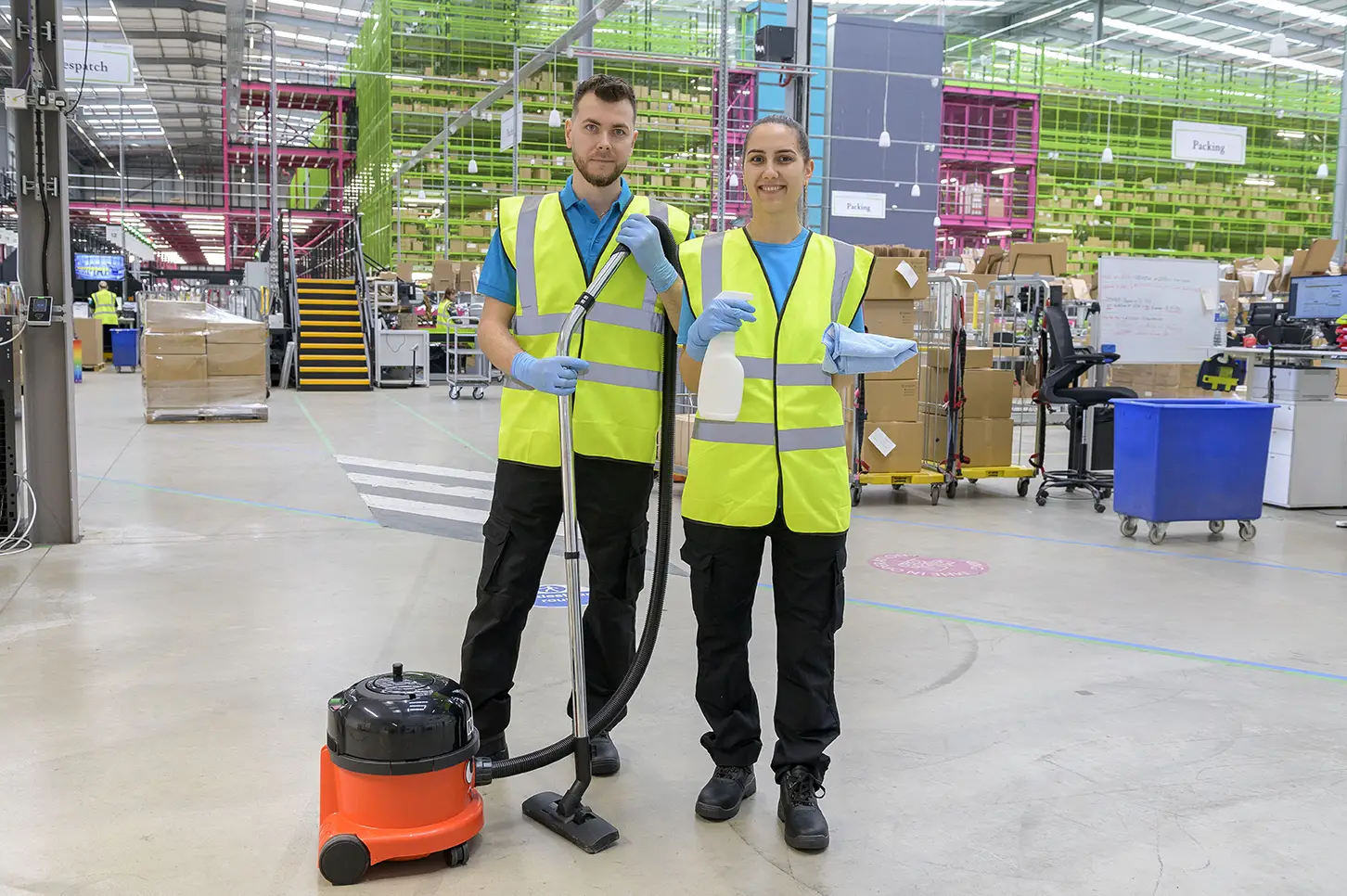 Two cleaners in branded polos and hi-vis vests with a vacuum, spray bottle and cloth in a warehouse
