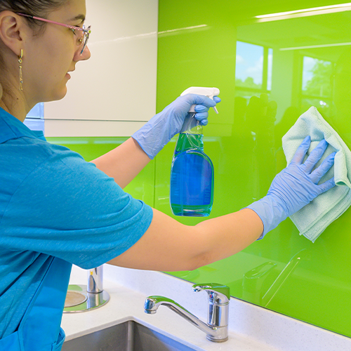 A cleaner in branded blue polo spraying and wiping a glossy green kitchen splashback above a sink