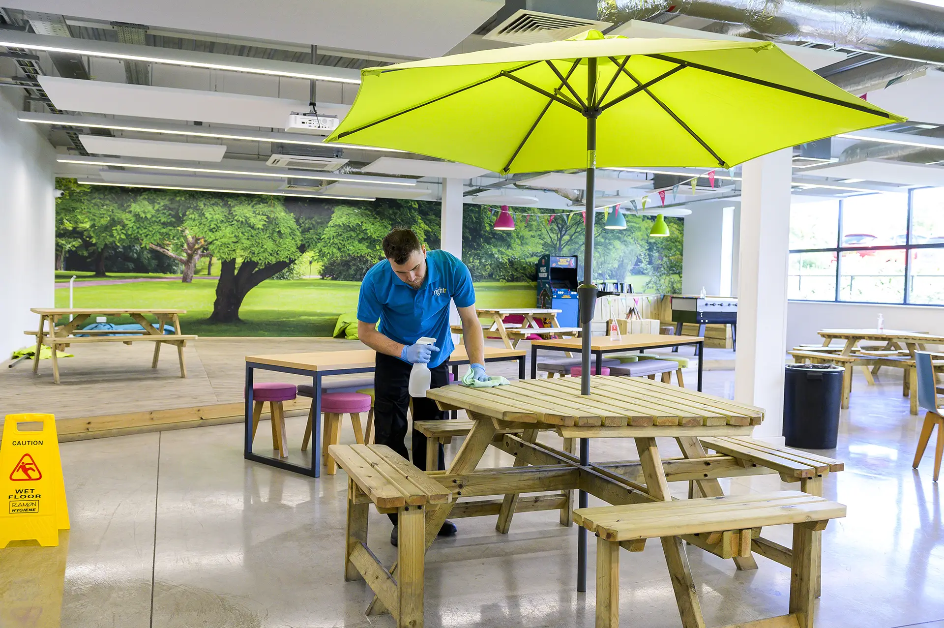 A cleaner in branded polo spraying a wooden picnic table indoors, under a yellow parasol