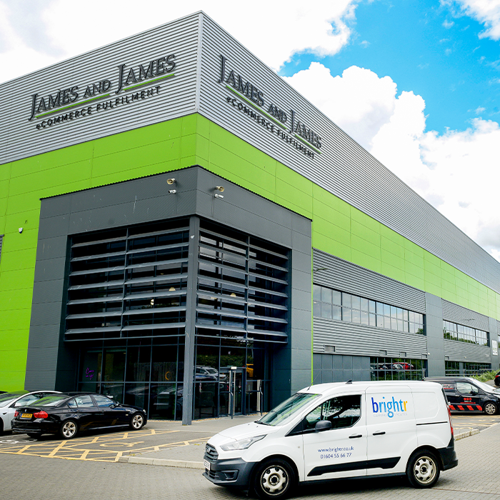 A large warehouse building with company signage, a branded van and cars parked outside