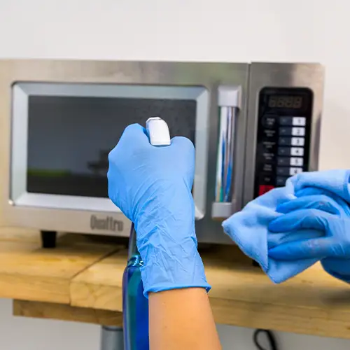 Gloved hands spraying and wiping down a stainless steel commercial microwave with a blue cloth