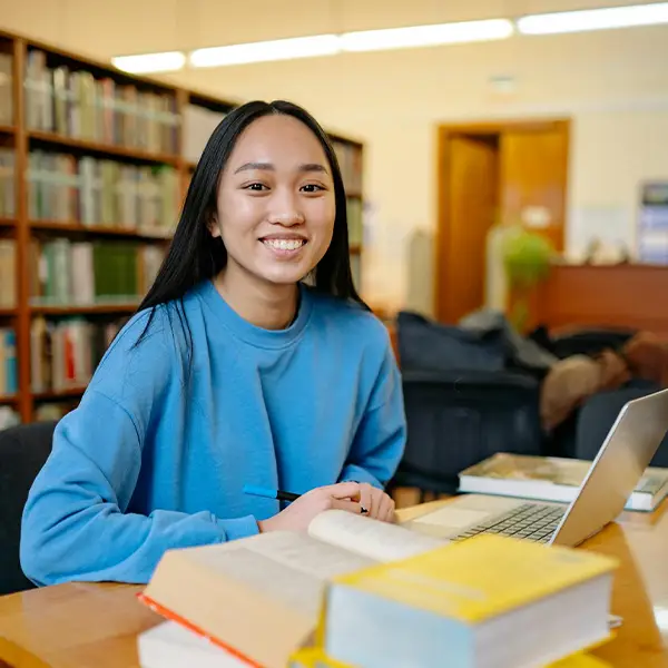 A young woman smiling at a desk with a laptop and stacked books, bookshelves behind her