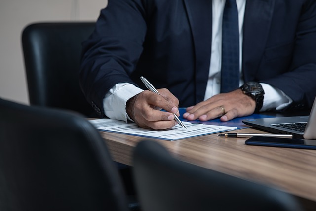 A person in a suit signing a document at a desk beside a laptop and pen