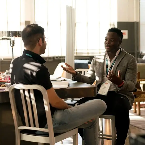 Two people talking at a table in a bright room with tall windows, one wearing a lanyard