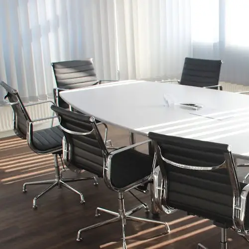 An empty boardroom with black leather chairs around a white table and vertical blinds at the window