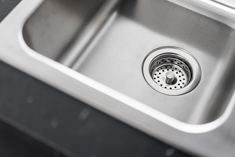 A close-up of a clean stainless steel sink with drain strainer, dark worktop edge