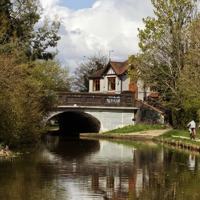 A stone bridge over a canal with a house behind it, trees along the bank and a cyclist on the towpath