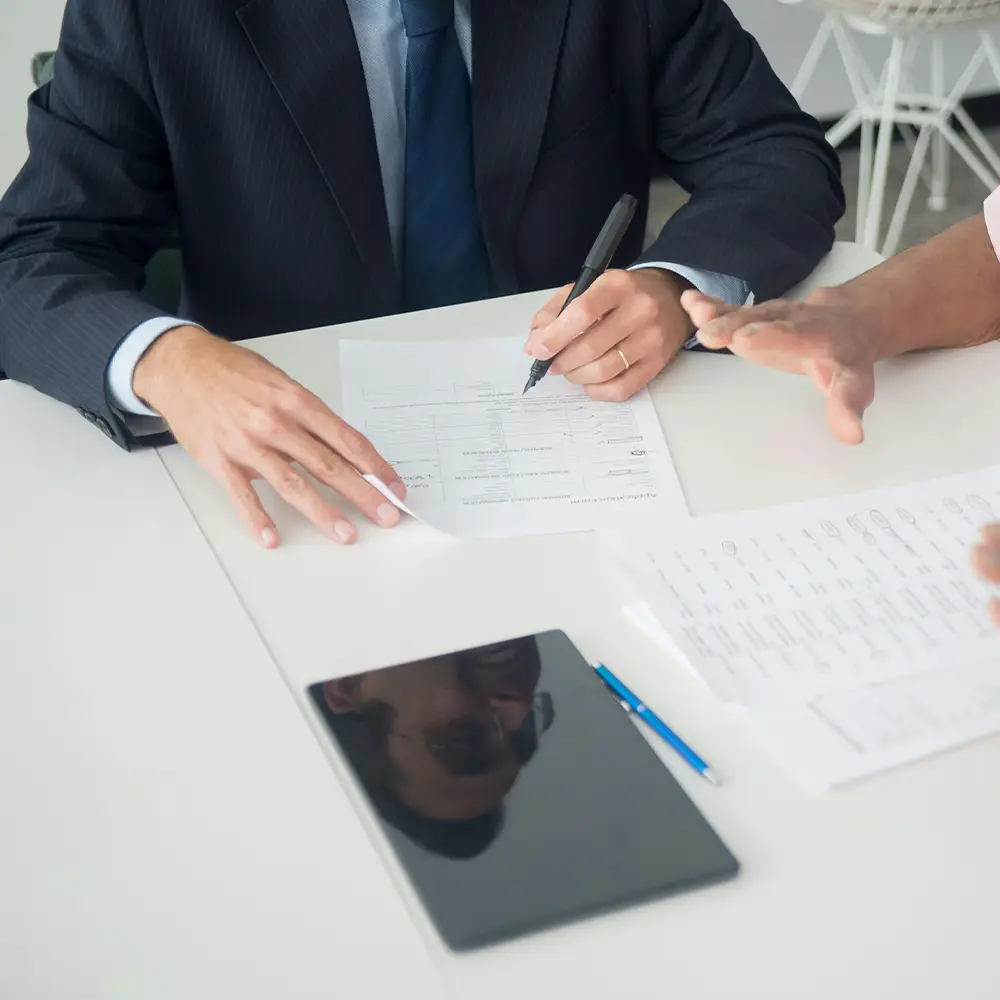 A person in a suit signing a document at a desk while another person gestures across the table