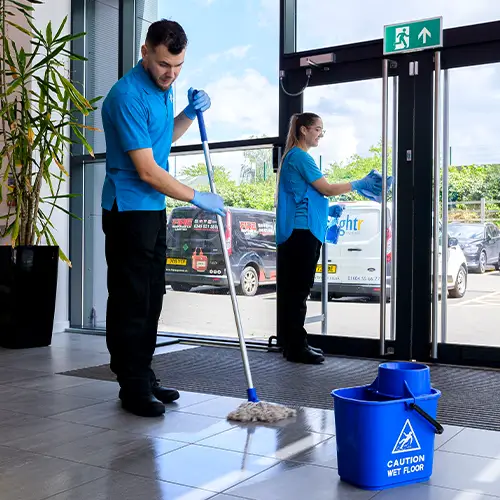 Two cleaners in branded uniforms mopping a glazed entrance, with a wet floor sign and branded van parked outside