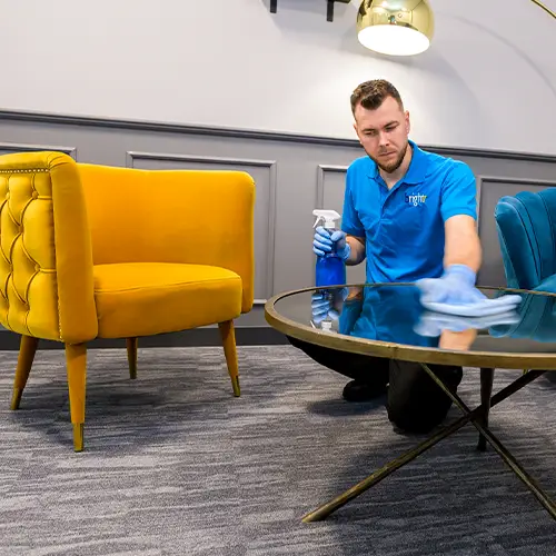 A cleaner in branded blue polo shirt and blue gloves wiping a glass coffee table beside a yellow armchair