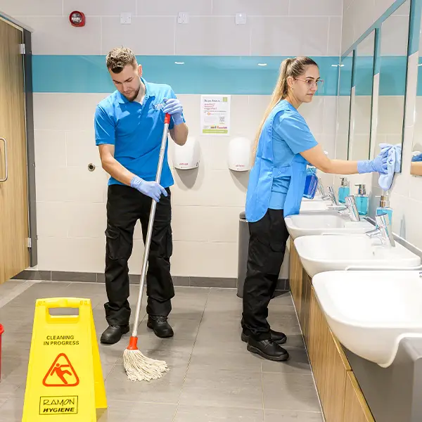 Two cleaners in a washroom, one mopping by a wet floor sign, the other wiping a mirror