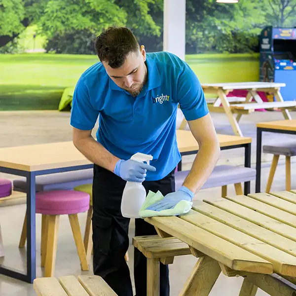 A cleaner in a branded polo wiping a wooden picnic table with a cloth and spray bottle, mural backdrop