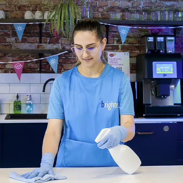 A cleaner in a branded apron spraying a white counter with a cloth, a coffee machine and bunting behind
