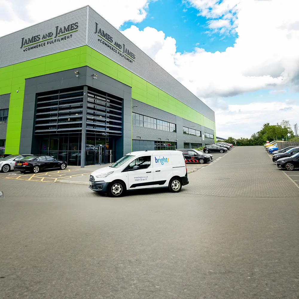 A large warehouse with green and grey cladding, a branded van and cars in a car park