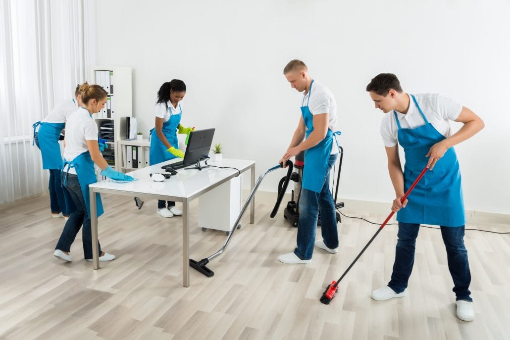 A team of cleaners in aprons vacuuming, mopping and wiping desks in an open-plan office