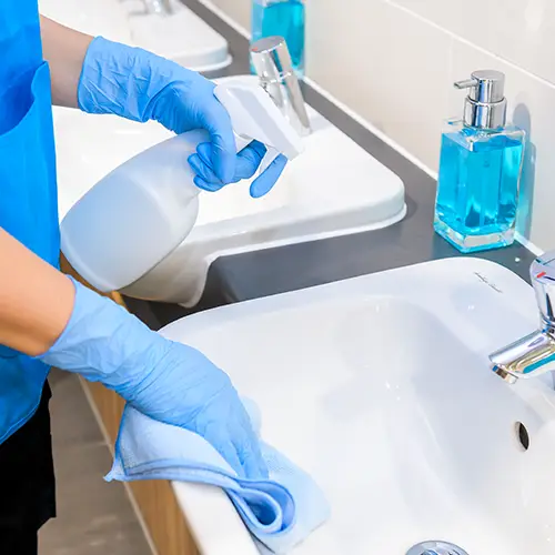 A cleaner in blue gloves spraying and wiping a bathroom sink beside soap dispensers