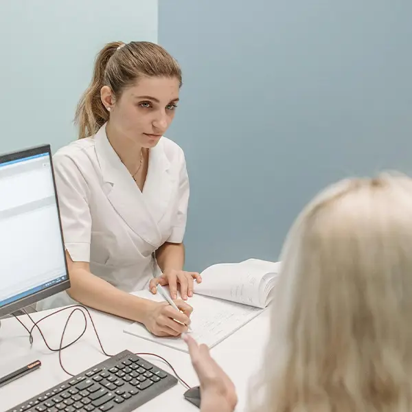 A person in a white coat at a desk with a computer, talking to someone across the desk
