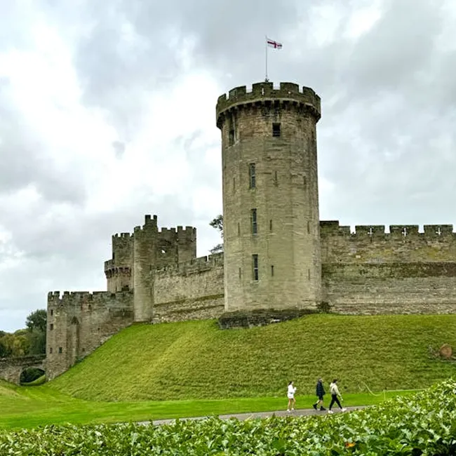 A stone castle turret with battlements and a flag, people walking on a path below
