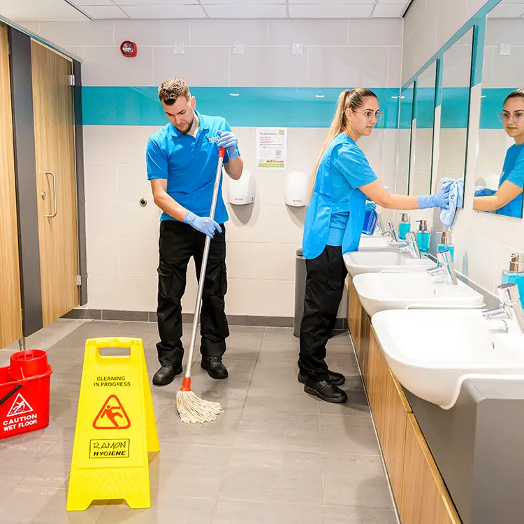 Two cleaners in branded uniforms mopping the floor and wiping mirrors in a washroom with a row of sinks