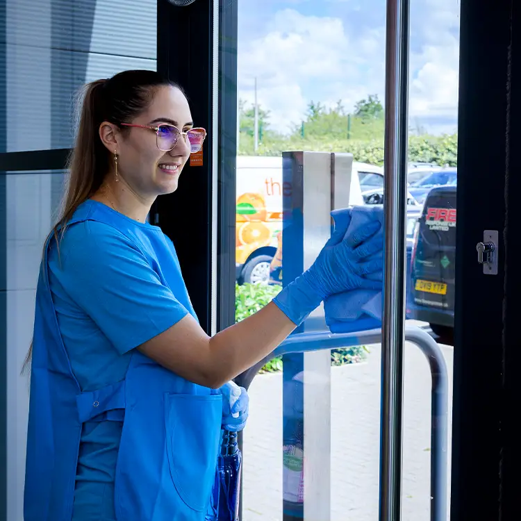 A cleaner in branded blue uniform and blue gloves polishing a stainless steel door frame at a glazed entrance