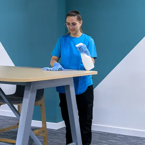 A cleaner in branded polo and blue gloves spraying and wiping a wooden table in an office space